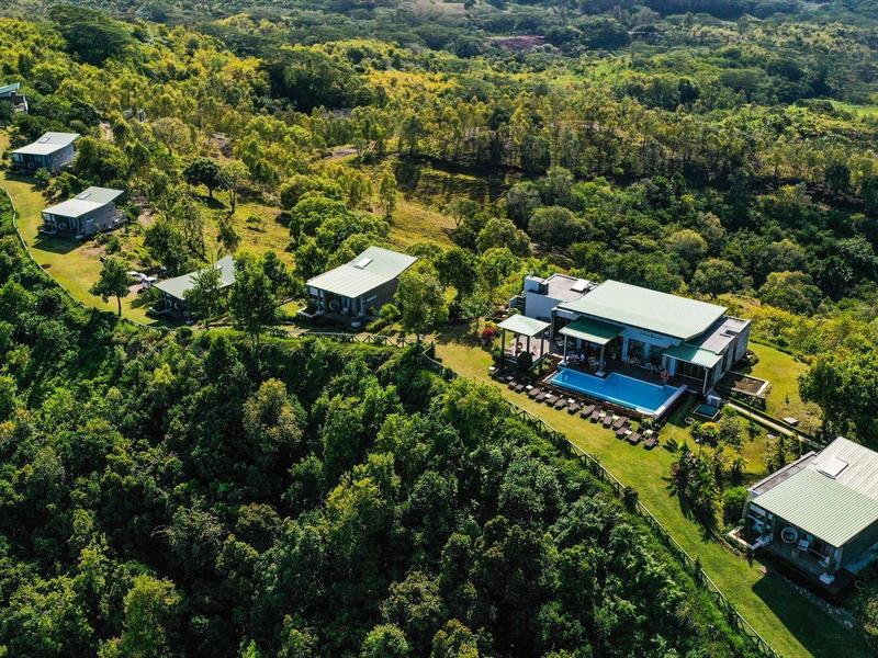 Luftaufnahme mehrerer moderner Häuser auf grünem Hügel, umgeben von dichtem Wald und Natur.