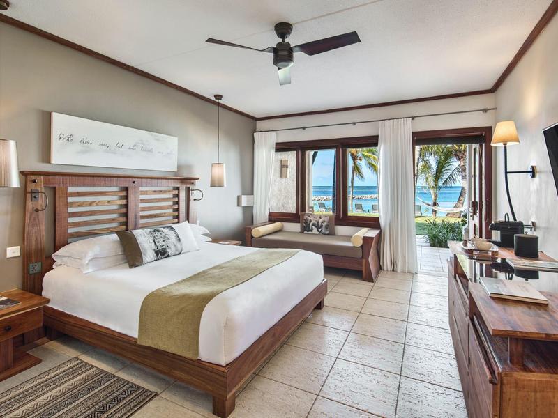 Hell eingerichtetes Hotelzimmer mit Doppelbett und Blick auf tropische Bäume und Meer.