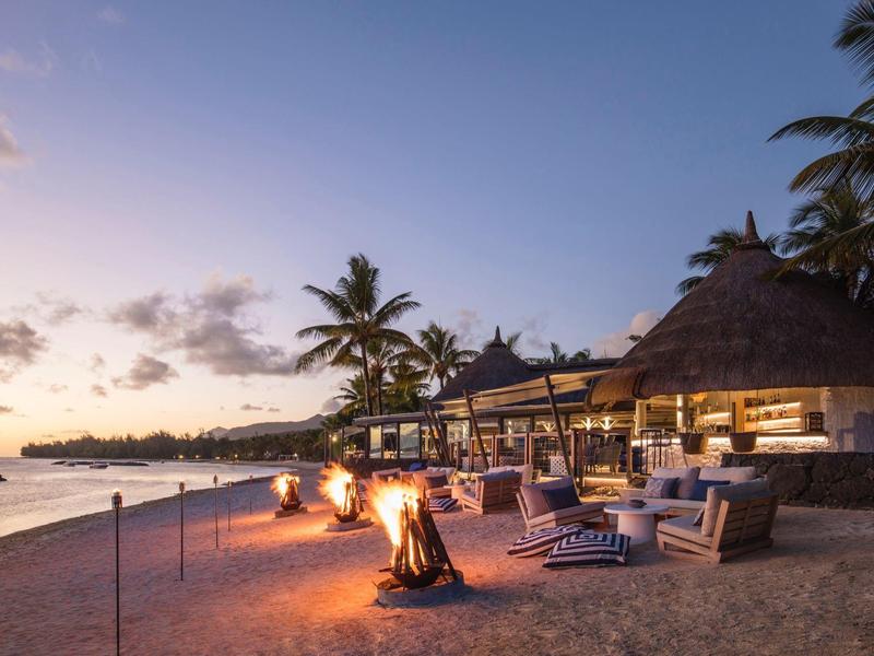 Strand mit Palmen, Lagerfeuer und gemütlichen Sitzecken bei Sonnenuntergang am Meer.