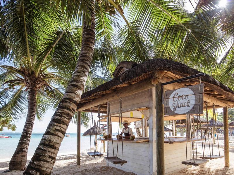 Kleine Strandbar mit Strohdach zwischen Palmen vor weißem Sand und blauem Meer.