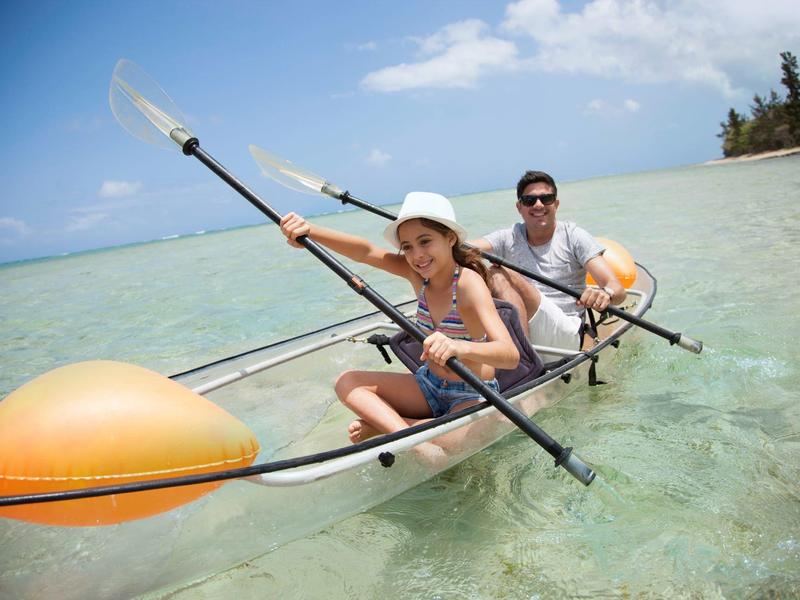 Zwei Personen paddeln in einem durchsichtigen Kajak auf klarem, flachem Wasser bei Sonnenschein.