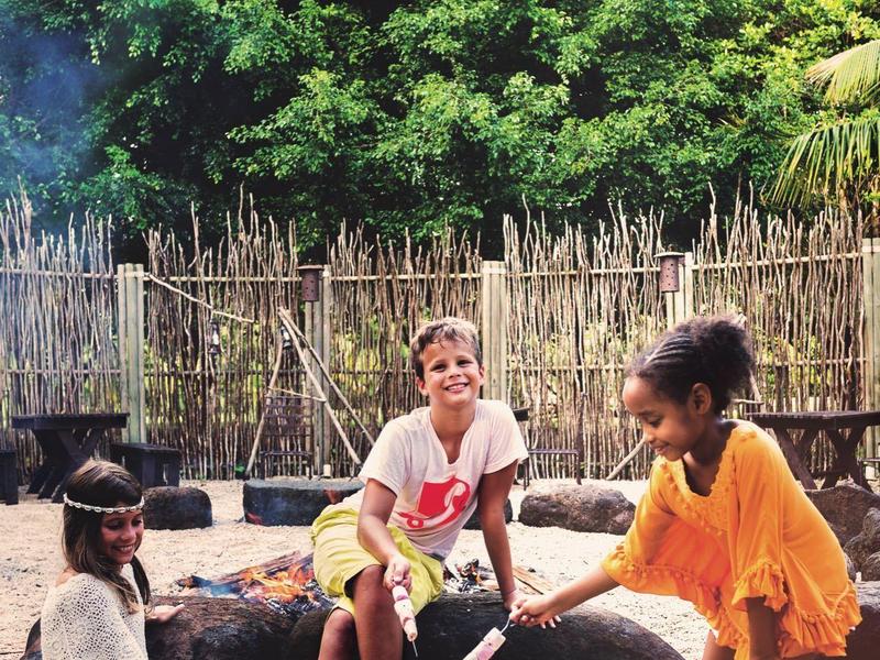 Drei Kinder sitzen und stehen draußen, kochen über einem kleinen Holzfeuer vor einem Zaun aus Ästen.