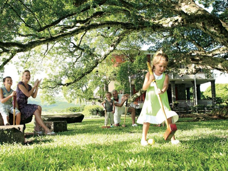 Familie spielt im Garten bei Sonnenschein, grüner Rasen und großer Baum mit ausladenden Ästen.