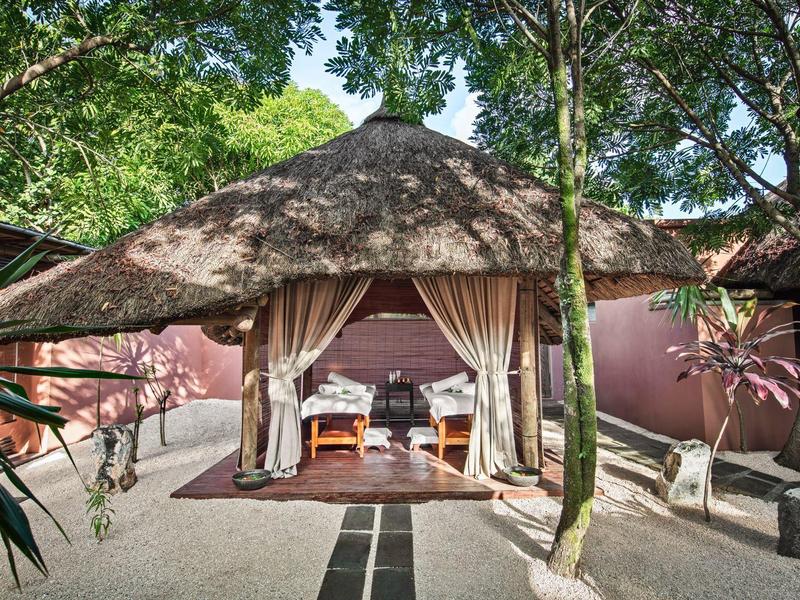 Offener Pavillon mit Strohdach, Vorhängen, Möbeln und Pflanzen in einem grünen Gartenbereich.