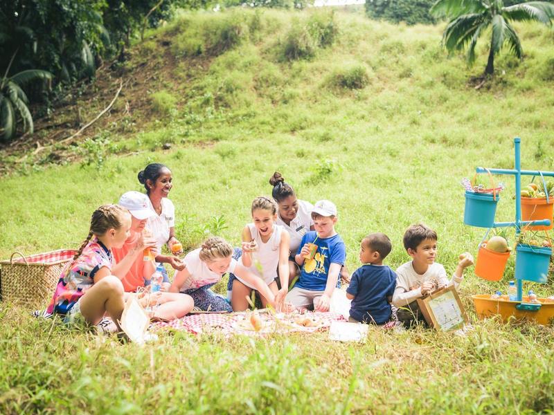 Eine Gruppe Kinder sitzt auf einer Wiese, neben ihnen stehen bunte Eimer auf einem Gestell.