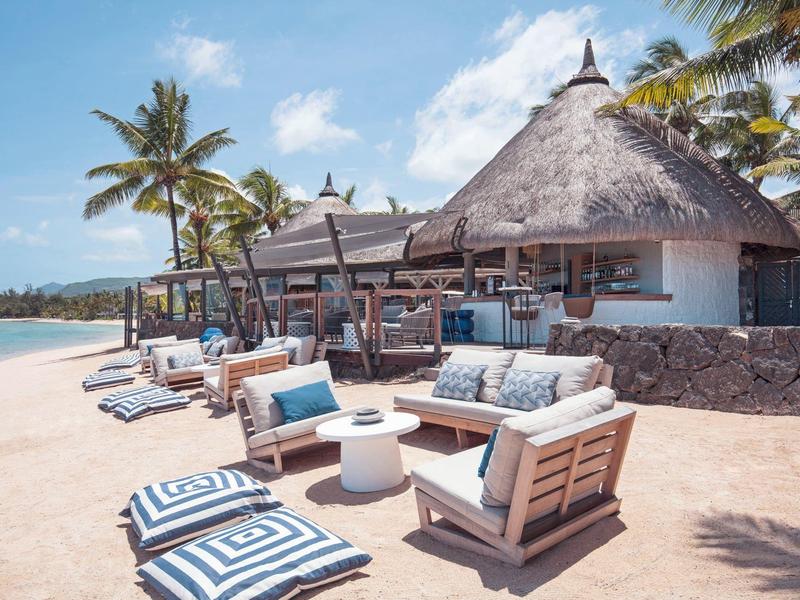 Strand mit weißen Sand, Palmen und gemütlichen Sitzmöbeln unter Strohdächern bei klarem Himmel.