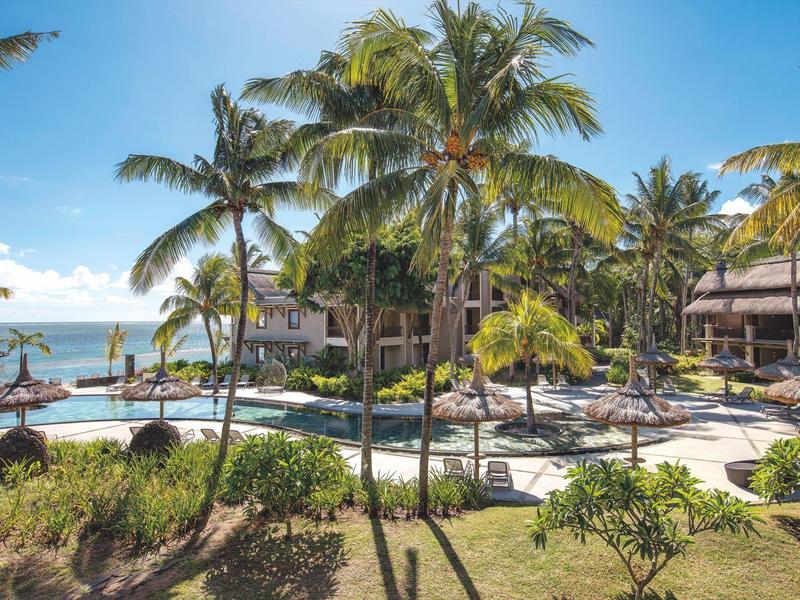 Tropische Poollandschaft mit Palmen, Liegestühlen und Bungalows am Meer bei sonnigem Wetter.