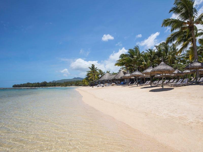 Weißer Sandstrand mit Palmen, Sonnenschirmen und klarem, ruhigem Wasser unter blauem Himmel.