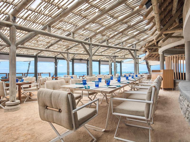 Offene Terrasse mit Sandboden, weißen Tischen, blauen Gläsern und Möwenblick am Meer.