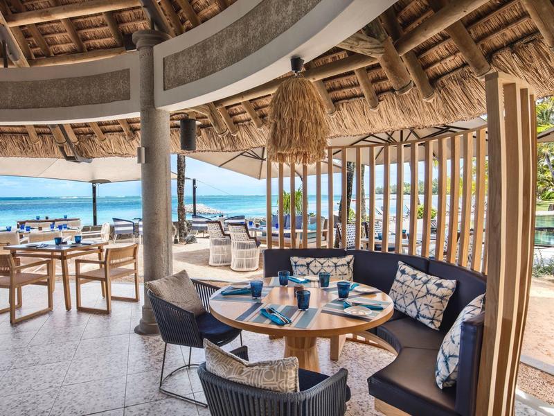 Offener Strandrestaurantbereich mit runden Tischen, Korbsesseln, Holzdecke und Blick auf das Meer.