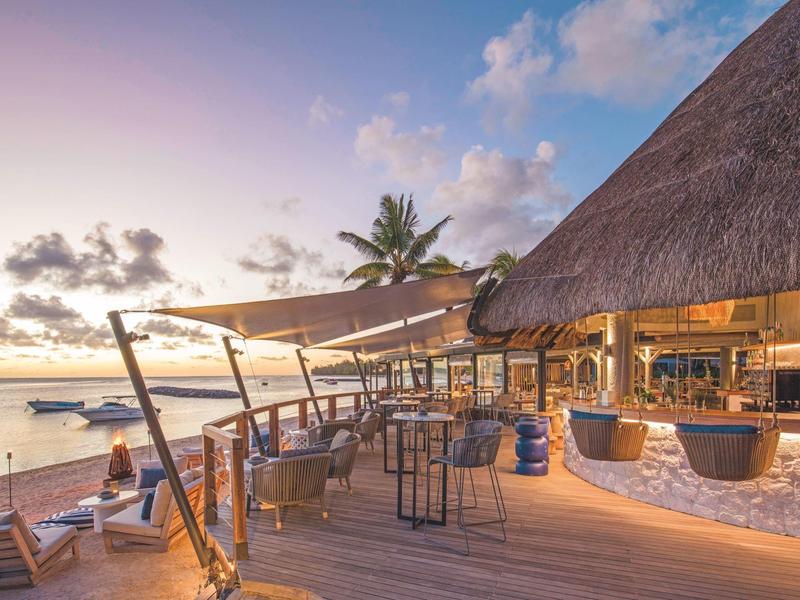 Strandbar mit Holzterrasse, Stühlen, Sonnensegel, Palmen und Booten im Sonnenuntergang.
