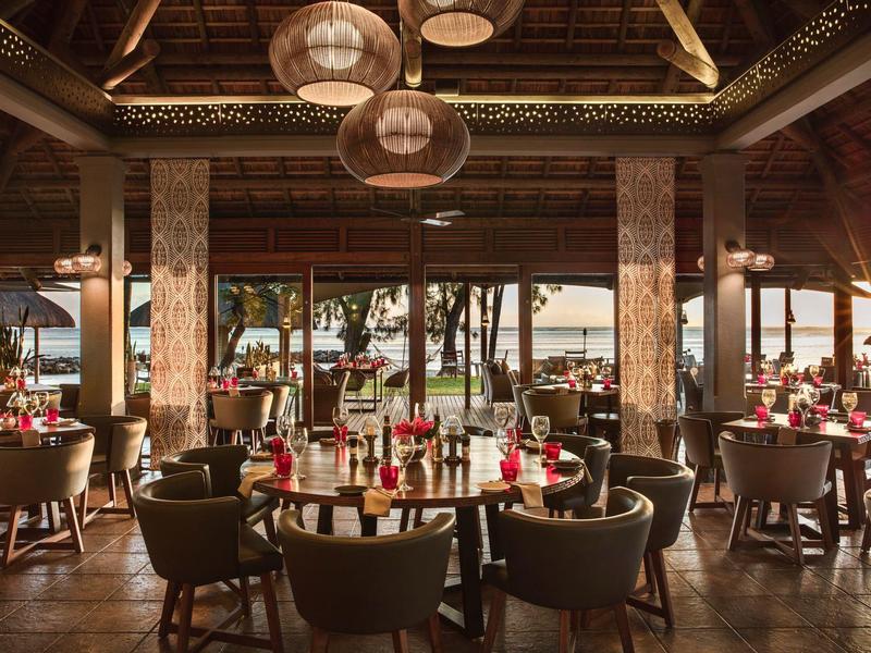 Offenes Restaurant mit Holzdach, runden Tischen, Stühlen und roter Tischdekoration am Strand.