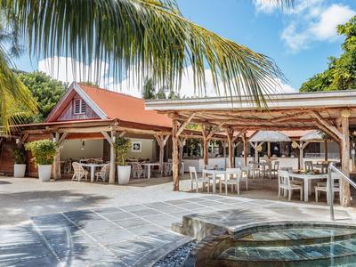 Freiluftrestaurant mit Holzdach, weißen Tischen und Stühlen, Palmen und Pool im Vordergrund.