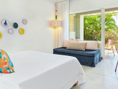 Jasny pokój hotelowy z podwójnym łóżkiem, kolorową poduszką, sofą i widokiem na taras z roślinami.