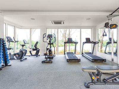 Heller Fitnessraum mit Sportgeräten und großen Fenstern, grau-blauer Teppich, weiße Wände.