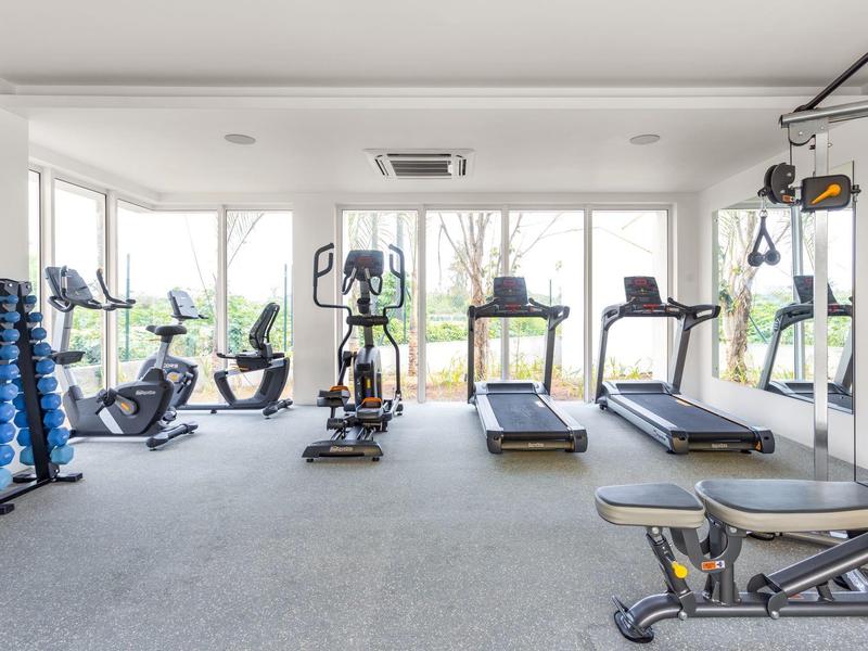 Brightly lit fitness room with treadmills, bikes, and dumbbells near large windows.
