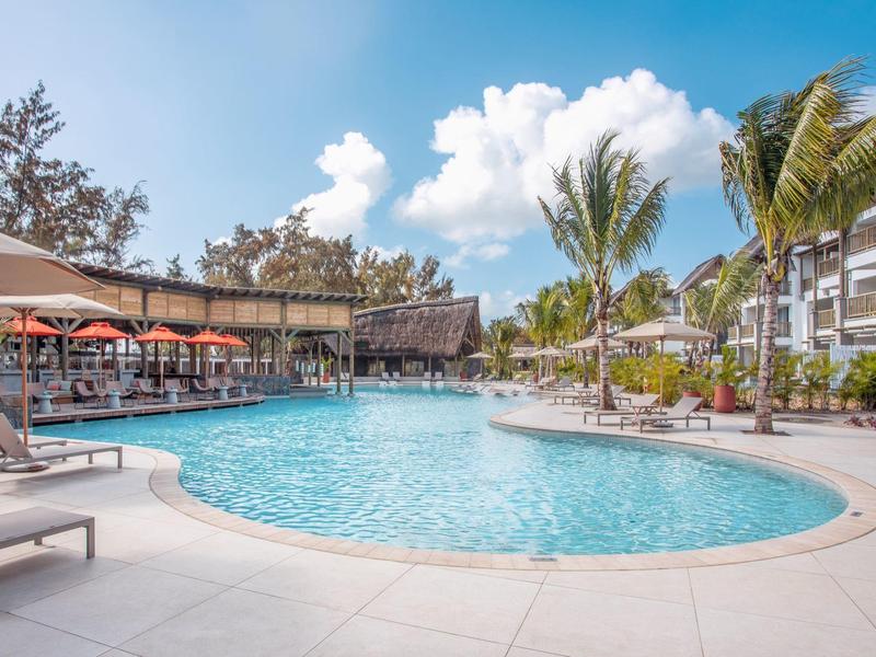 Large hotel resort pool area with lounge chairs and palm trees under a blue sky.