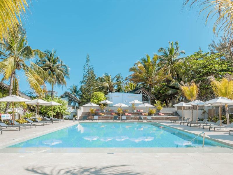Large pool with lounge chairs and umbrellas surrounded by palm trees under a blue sky.