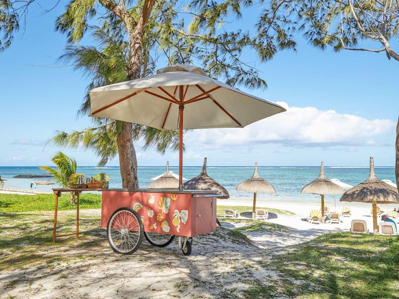 Colorful ice cream cart under a parasol on the beach with loungers and sea in the background.