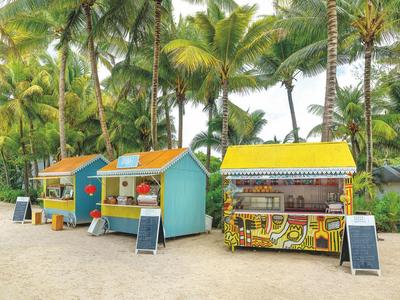 Drei bunte Marktstände am Strand, umgeben von hohen Palmen und Sandboden, unter hellem Himmel.