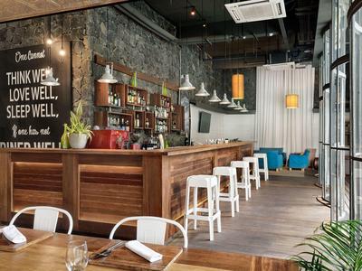 Modernes Café mit Holztheke, weißen Barhockern, beleuchteten Regalen und Betonwand mit Schriftzug.