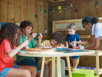 Děti si hrají a jedí společně u kulatého dřevěného stolu v útulné místnosti.