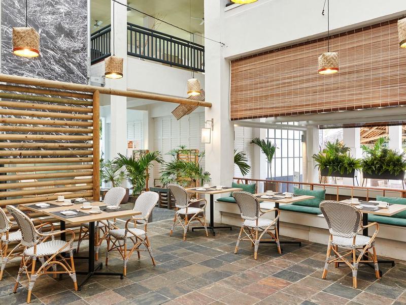 Moderní restaurace se světlým nábytkem, dřevěnými akcenty a rostlinami, zalitá sluncem.