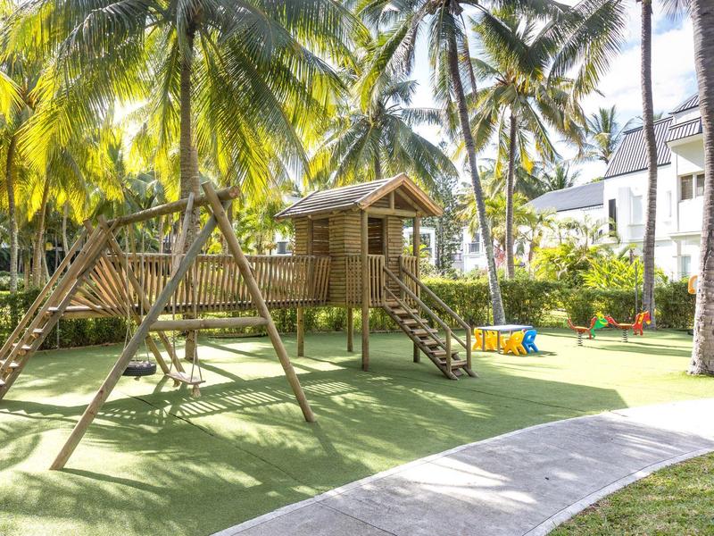 Dětské hřiště s dřevěným domkem a houpačkami v tropickém hotelu s palmami