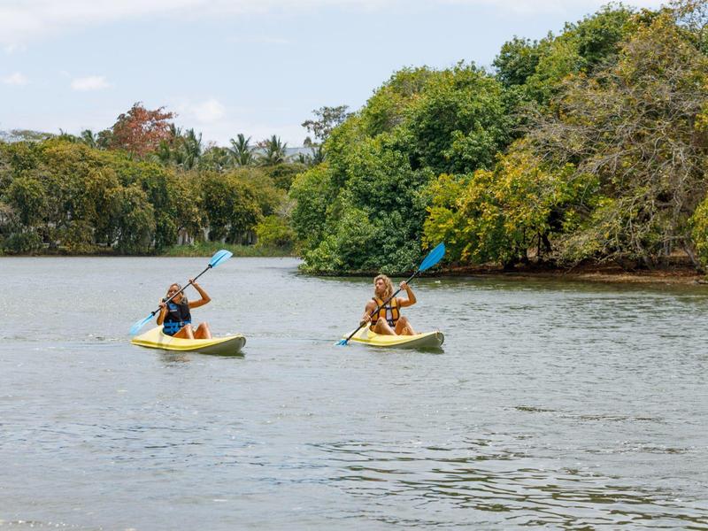 Dva lidé pádlí v žlutých kajacích na klidné řece se zelenými břehy.