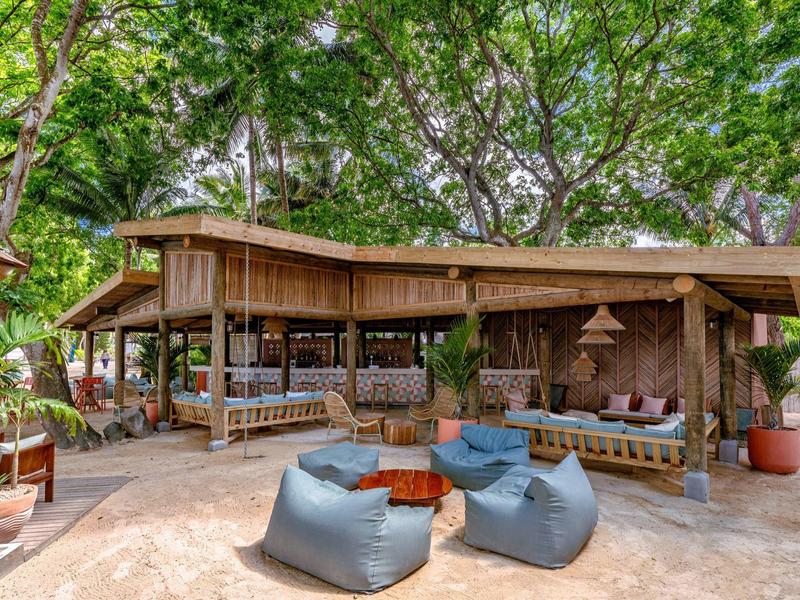 Otevřený lounge s vaky na sezení a dřevěným nábytkem pod hustou zelení.