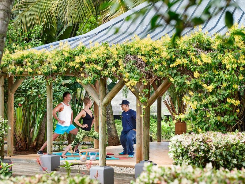 Tři lidé na dlážděné cestě pod květinově zdobenou dřevěnou pergolou v tropické zahradě.