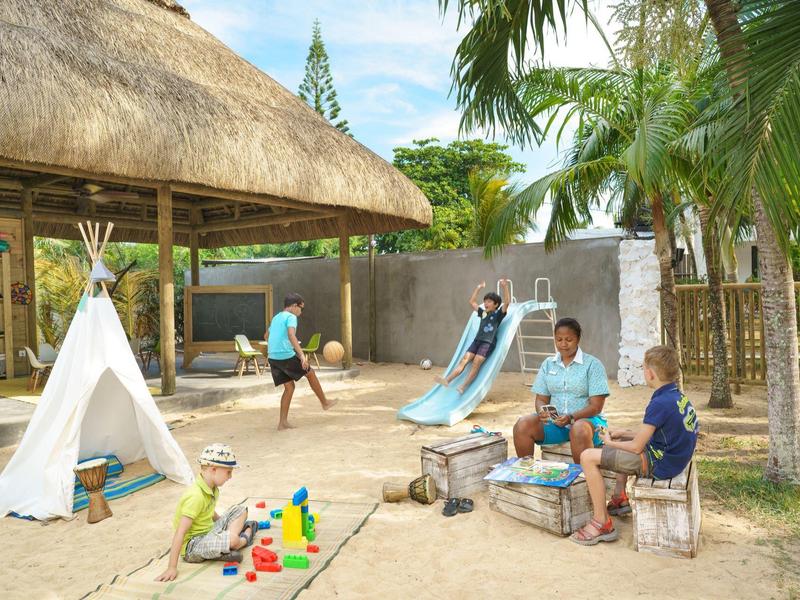 Kinder spielen auf einem Sandspielplatz mit Rutsche und Zelt in einem tropischen Garten.