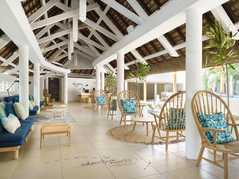 Offener Hotel-Loungebereich mit weißen Balken, Korbstühlen und blauen Kissen.