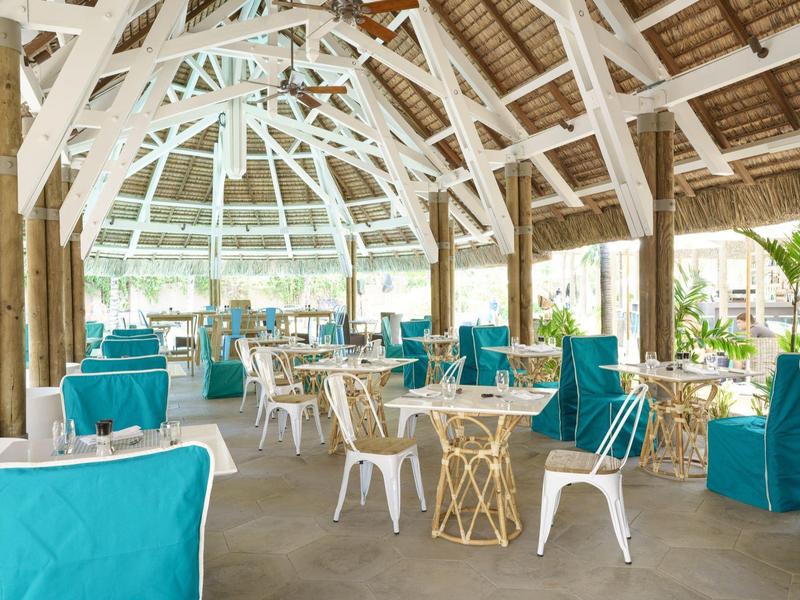 Offener, luftiger Restaurantbereich mit blauen Sitzkissen und Holzdach unter Palmen.