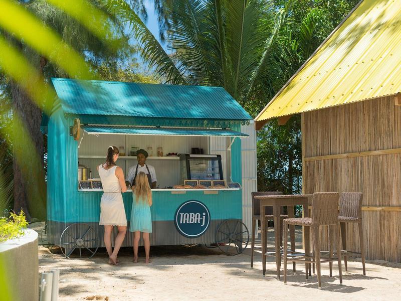 Zwei Frauen bestellen an einem kleinen blauen Strandkiosk neben einer Hütte mit Strohdach.