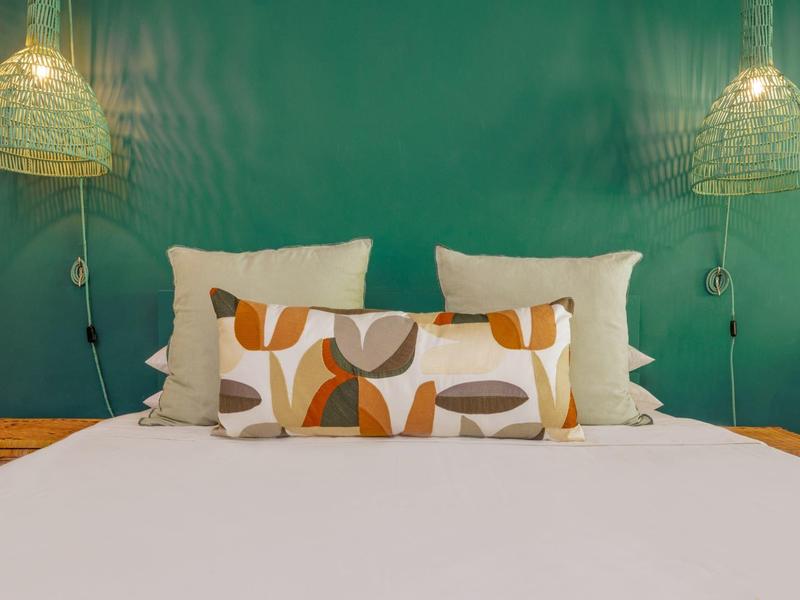 Gemütliches Bett mit weißen Kissen und dekorativem Kissen vor grünem Hintergrund und zwei Hängelampen