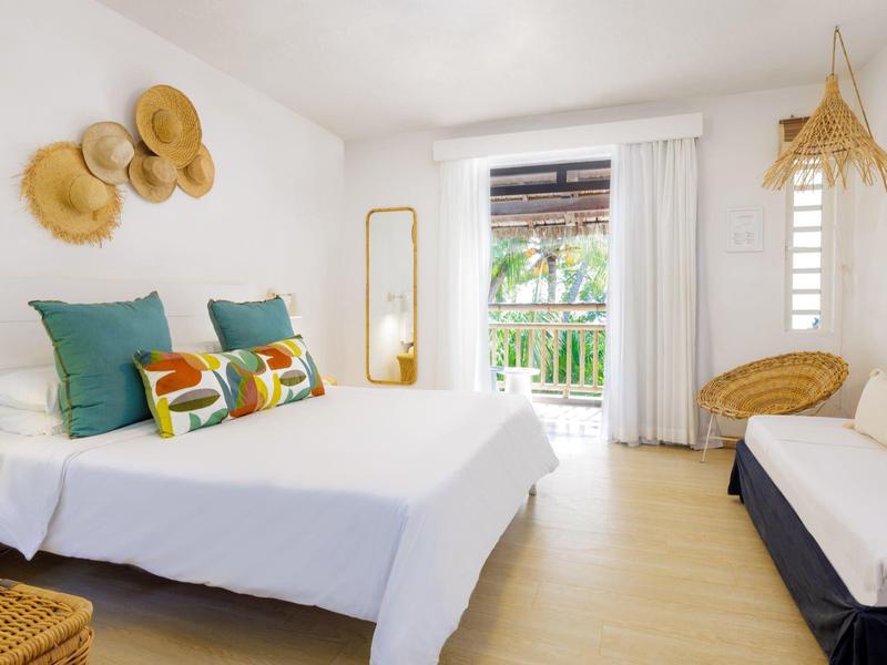 Helles Schlafzimmer mit weißer Bettwäsche, bunten Kissen und Balkon mit Pflanzenblick.