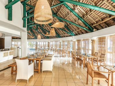 Světla restaurace s dřevěnými židlemi, velkými okny a tkanými stropními lampami.