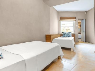 Světložlutá ložnice se dvěma jednolůžky, dřevěným nábytkem a oknem s žaluziemi.