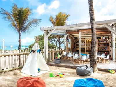 Pobřežní oblast s chatkou, palmami, stanem teepee a barevnými sedacími vaky pod modrou oblohou.