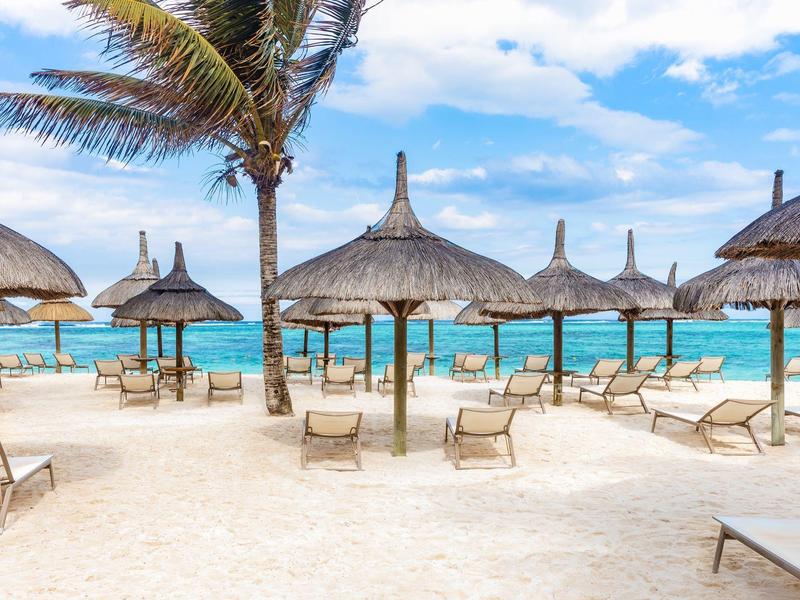 Pláž se slaměnými slunečníky a lehátky u tyrkysového moře pod modrou oblohou.