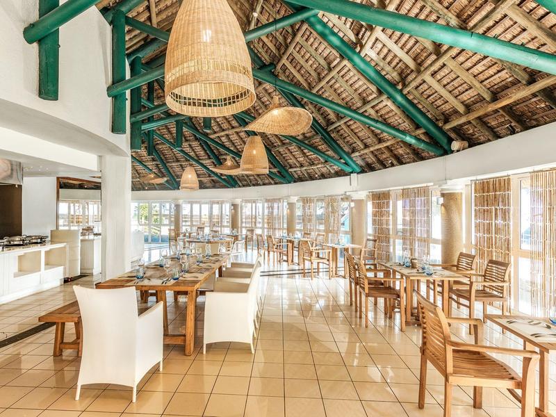 Světla restaurace s dřevěnými židlemi, velkými okny a tkanými stropními lampami.