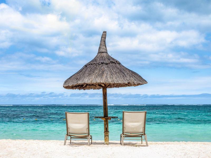 Dvě lehátka pod doškovou slunečníkem na bílé písečné pláži s modrým mořem a oblohou.