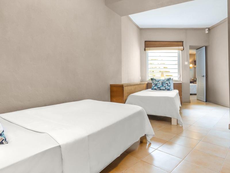 Světložlutá ložnice se dvěma jednolůžky, dřevěným nábytkem a oknem s žaluziemi.