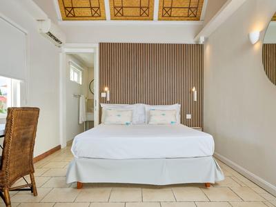 Světlý hotelový pokoj s velkou postelí, psacím stolem a židlí pod dekorativním stropem.