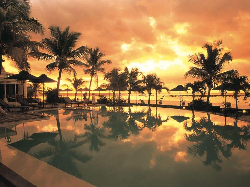 Západ slunce nad bazénem s palmami a lehátky na pláži hotelu.