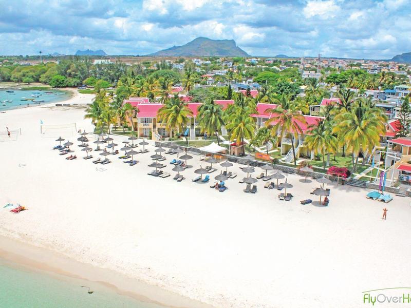 Pláž s deštníky a palmami před pobřežním městem a horami v pozadí.