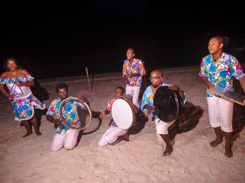 Skupina hudebníků na pláži v noci s tradičními bubny a barevnými košilemi.