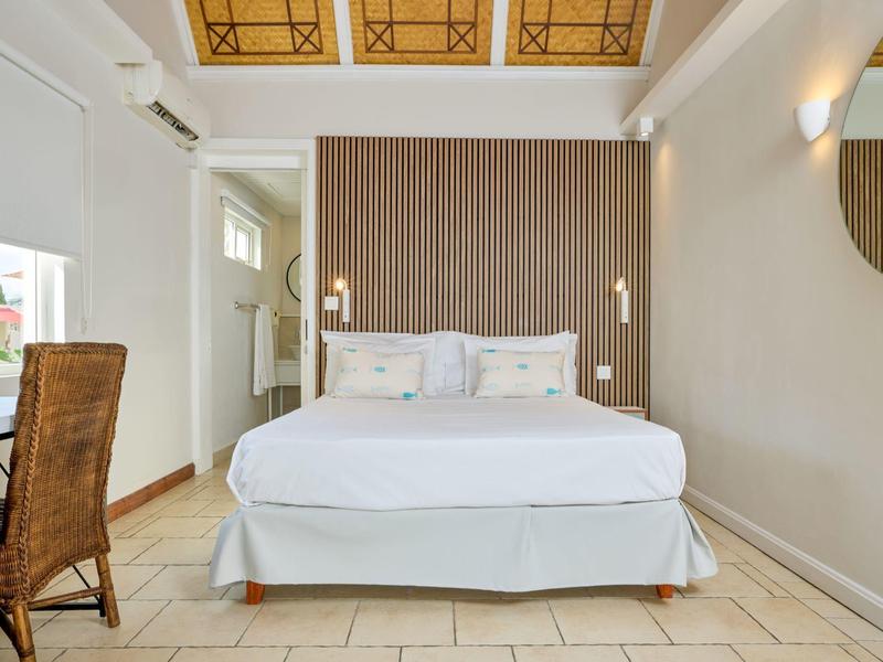 Světlý hotelový pokoj s velkou postelí, psacím stolem a židlí pod dekorativním stropem.