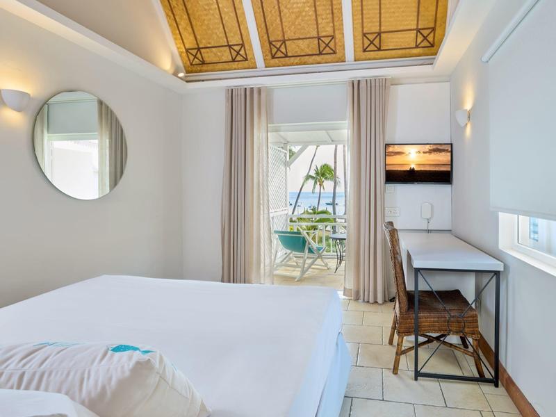 Světý hotelový pokoj s manželskou postelí, stolem, židlí a balkonem s výhledem na moře.